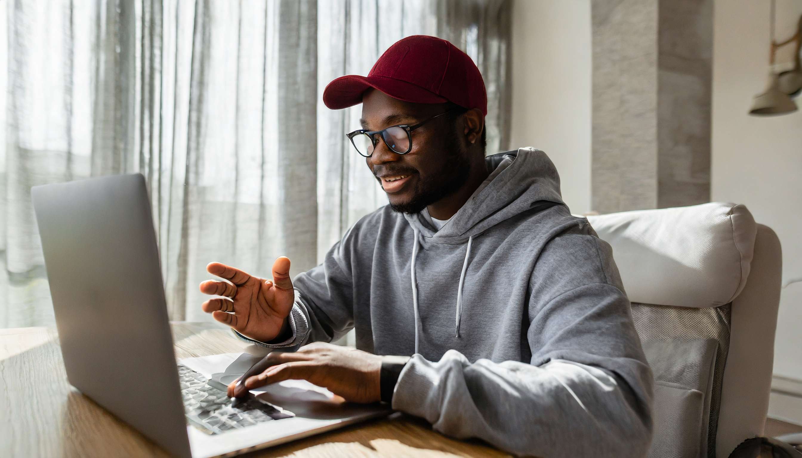 Firefly A young man with glasses in a maroon baseball cap and grey hoodie seated at a desk in their 